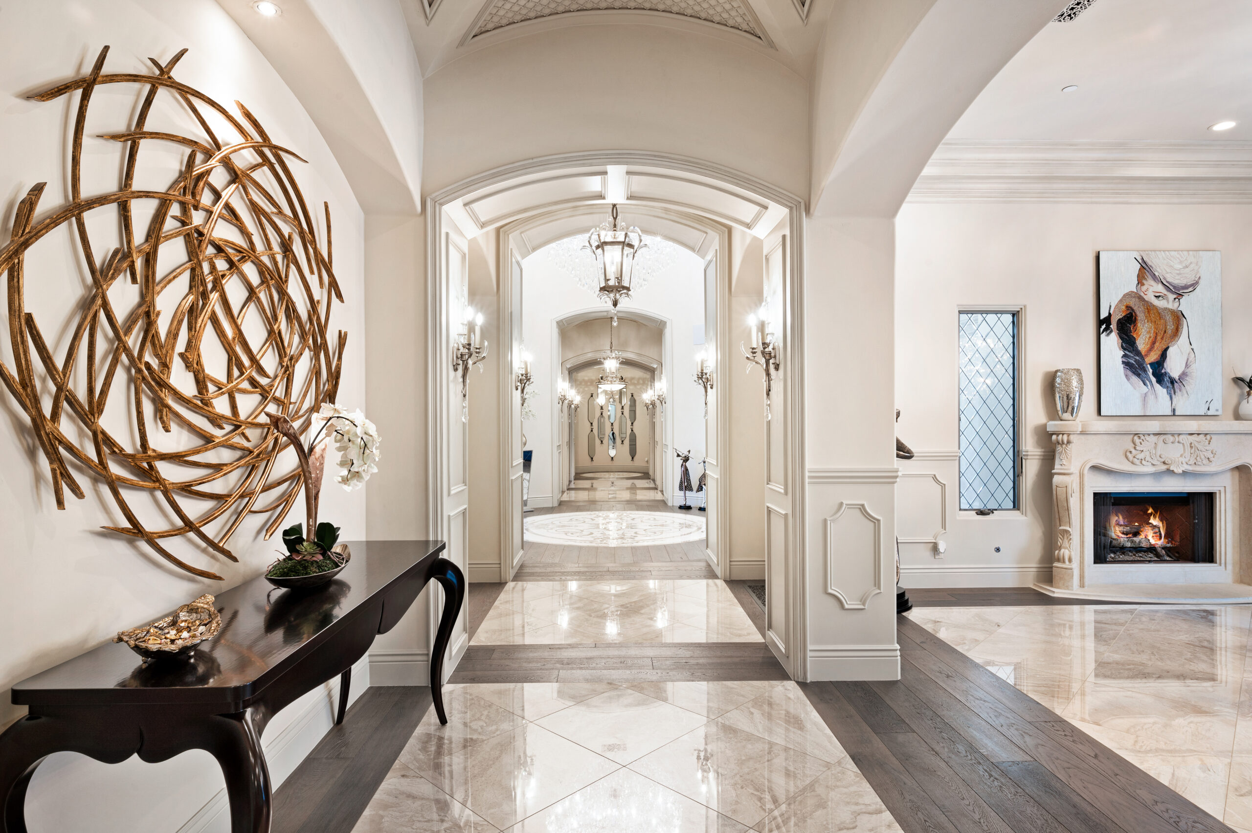 An elegant hallway featuring a stunning gold abstract wall sculpture on the left wall, adding a modern artistic touch. A dark wood console table beneath the sculpture is adorned with decorative items and a small potted plant. The hallway leads to a grand archway, highlighting the luxurious design with marble flooring and soft lighting from wall sconces and ceiling fixtures. In the background, an open space reveals a cozy living area with a fireplace, and additional artwork, creating a warm and inviting atmosphere.