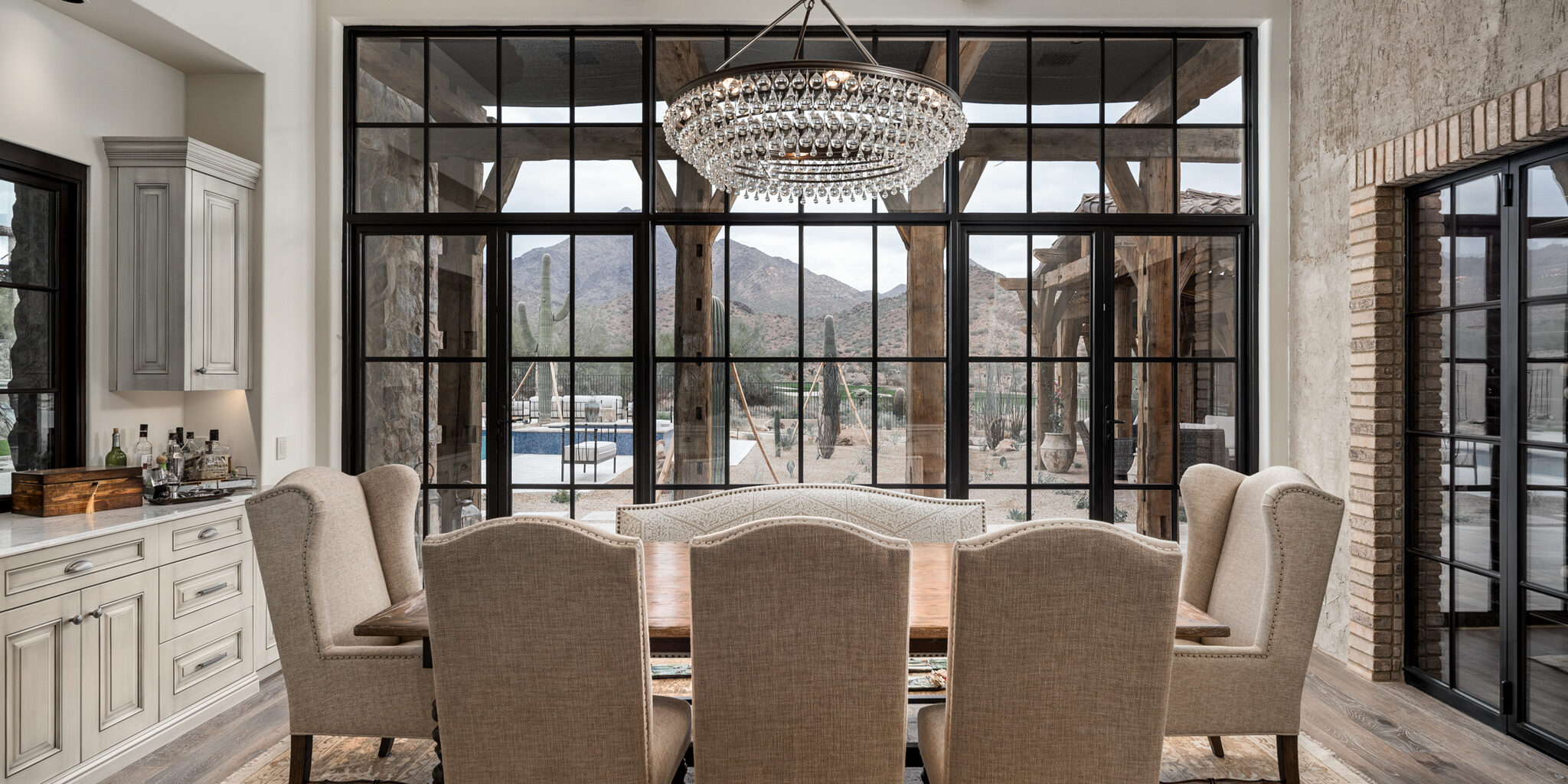 A beautifully designed dining room featuring a large rectangular wooden table surrounded by upholstered high-back chairs. The room has a rustic charm with exposed wooden beams on the ceiling and a large, elegant chandelier hanging above the table. The back wall is composed of expansive floor-to-ceiling windows with black frames, providing a stunning view of the outdoor scenery and mountains. The room is further enhanced by a vintage-style area rug and light wood flooring. Adjacent to the dining area, built-in cabinetry provides additional storage and complements the overall aesthetic. The natural light and open layout create a warm and inviting atmosphere.