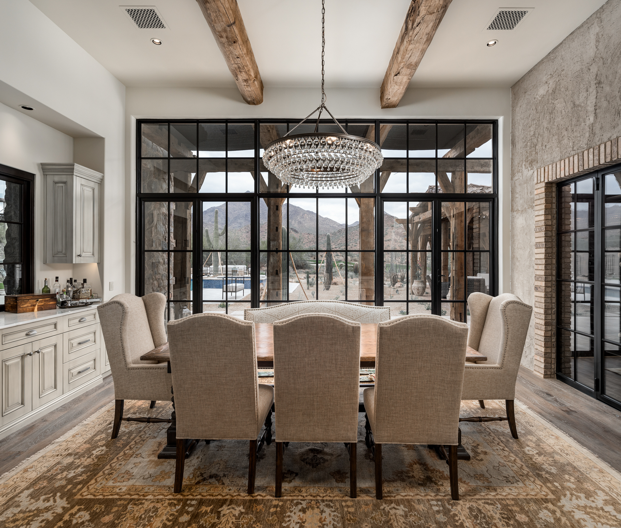 A beautifully designed dining room featuring a large rectangular wooden table surrounded by upholstered high-back chairs. The room has a rustic charm with exposed wooden beams on the ceiling and a large, elegant chandelier hanging above the table. The back wall is composed of expansive floor-to-ceiling windows with black frames, providing a stunning view of the outdoor scenery and mountains. The room is further enhanced by a vintage-style area rug and light wood flooring. Adjacent to the dining area, built-in cabinetry provides additional storage and complements the overall aesthetic. The natural light and open layout create a warm and inviting atmosphere.