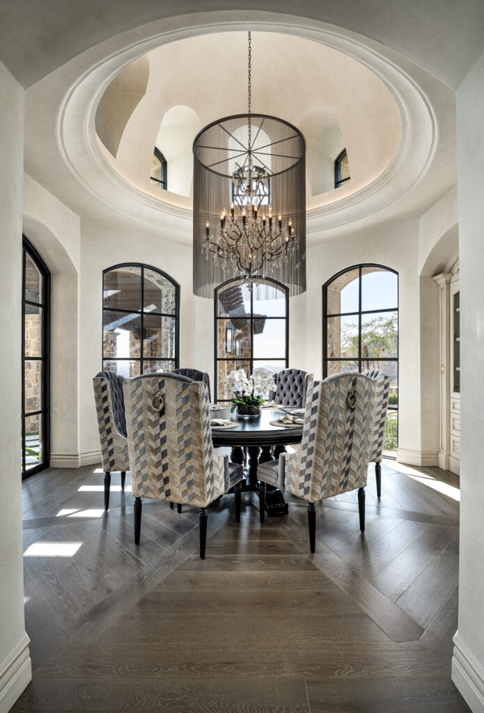 Dining Room with Chandelier and Arched Windows: A sophisticated dining room featuring a round dining table surrounded by high-backed patterned chairs. A large, decorative chandelier hangs from the ceiling, which has a domed design with recessed lighting. The room has multiple arched windows, allowing ample natural light.