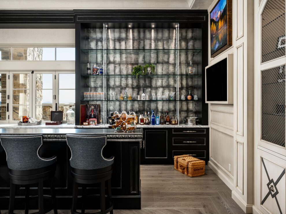 Close-Up of Home Bar Shelving and Counter: A close-up view of the home bar, highlighting the glass shelves displaying various bottles, a dark countertop, and bar stools. The backdrop is a textured grey wall with modern lighting, and there are cabinets with glass doors for additional storage.