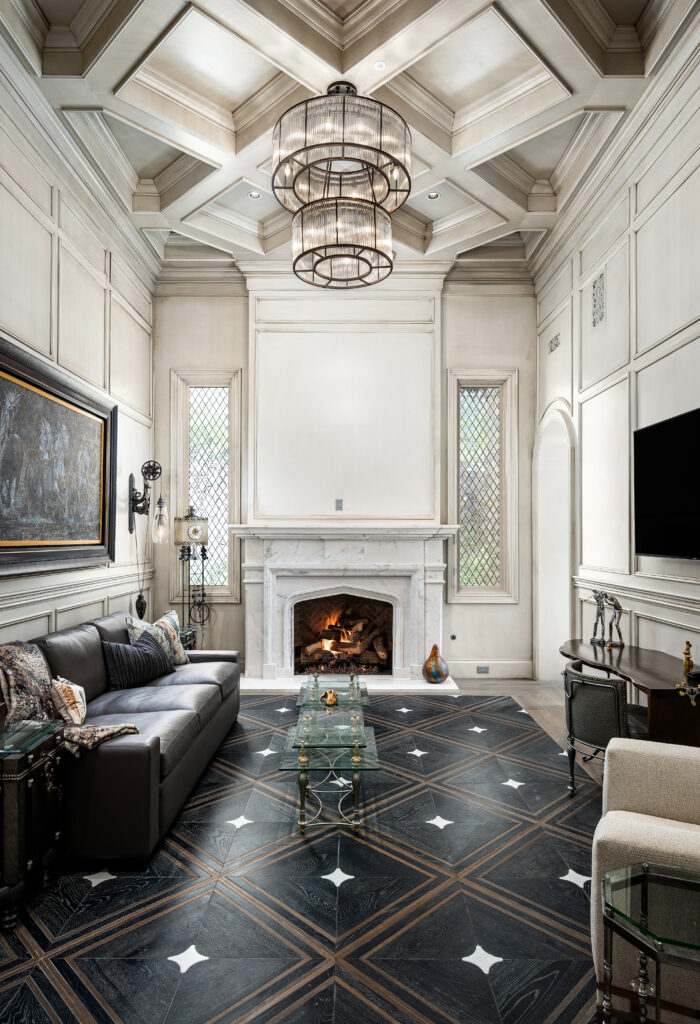 Sophisticated Living Room with Coffered Ceiling: A refined living room with a high coffered ceiling and a central chandelier. The room features a large marble fireplace, a black leather sofa, and a glass coffee table. The walls are adorned with paneling and decorative moldings, and tall windows with diamond-patterned glass allow natural light to fill the space.