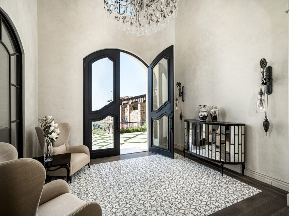 A luxurious foyer featuring a grand double door entrance with arched black frames that open to a scenic outdoor view. The room is elegantly decorated with a chandelier that hangs from the ceiling, casting a warm light over the space. The floor is adorned with a beautiful patterned tile, adding a touch of sophistication. On one side, there are comfortable beige chairs and a small table with a floral arrangement. On the other side, a stylish mirrored console table is decorated with vases and other decorative items. Wall sconces with hanging crystals add to the elegant ambiance of the foyer.