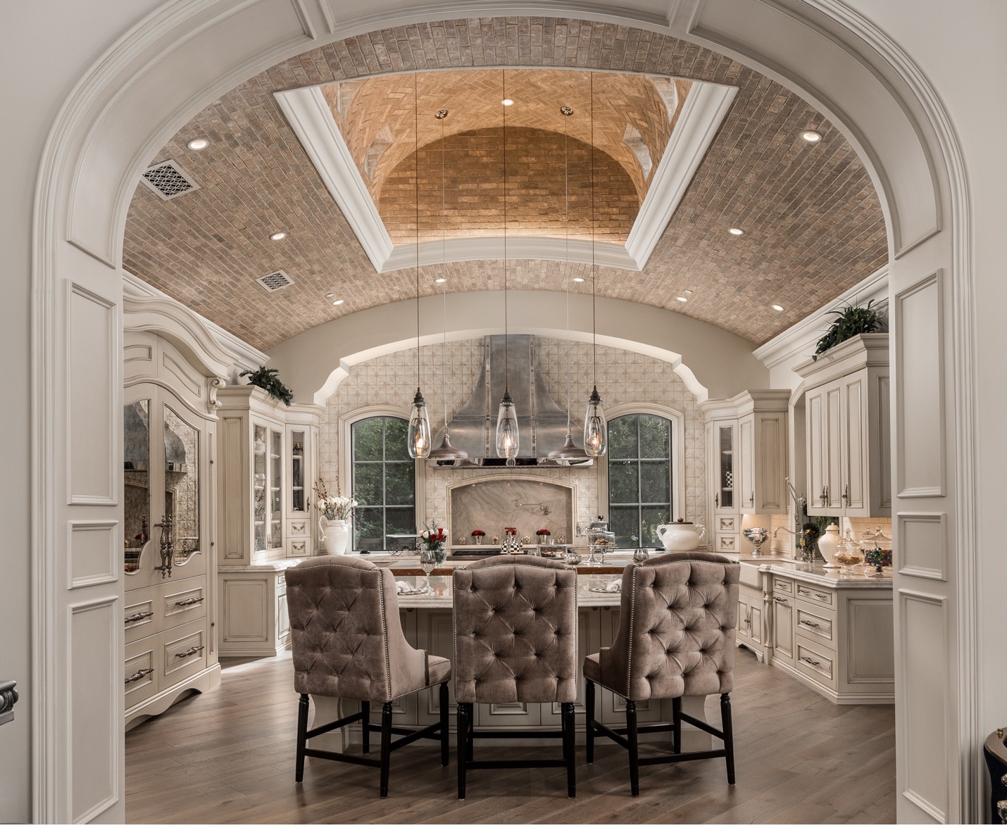 An exquisite kitchen featuring a high, arched brick ceiling with recessed lighting and a sophisticated pendant light fixture. The space is centered around a large island with a white marble countertop and tufted upholstered barstools. The cabinetry is painted in a soft cream color, complementing the elegant design. A grand range hood, flanked by windows, sits above the professional-grade stove. The kitchen is adorned with intricate moldings and detailed paneling, creating a classic and luxurious ambiance. The floor is covered with light wood planks, adding warmth to the refined decor. A decorative archway frames the view into this beautifully designed kitchen, enhancing its opulent feel.