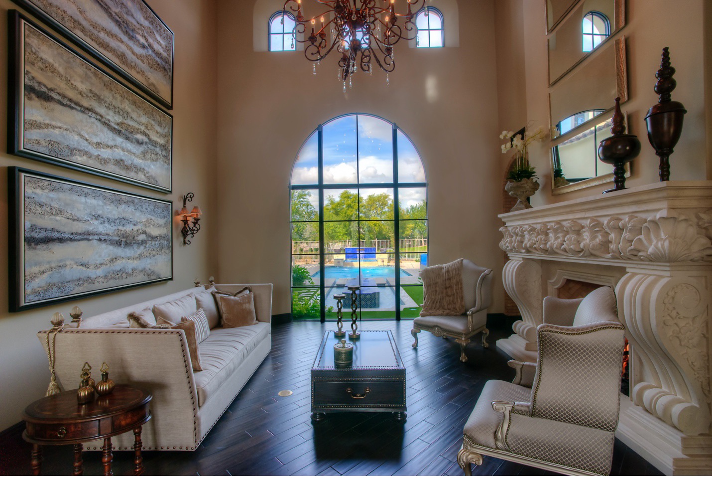 A luxurious Mediterranean-style living room featuring a high ceiling with elegant chandeliers and large arched windows offering a view of the outdoor pool area. The room is decorated with a grand, ornately carved fireplace mantel and a matching set of plush armchairs and a sofa in neutral tones. The walls are adorned with large abstract paintings and classic wall sconces. A dark wooden coffee table sits at the center of the room, complemented by a small side table with decorative items. The dark hardwood floors add warmth and contrast to the light-colored furnishings and walls, creating a sophisticated and inviting atmosphere.