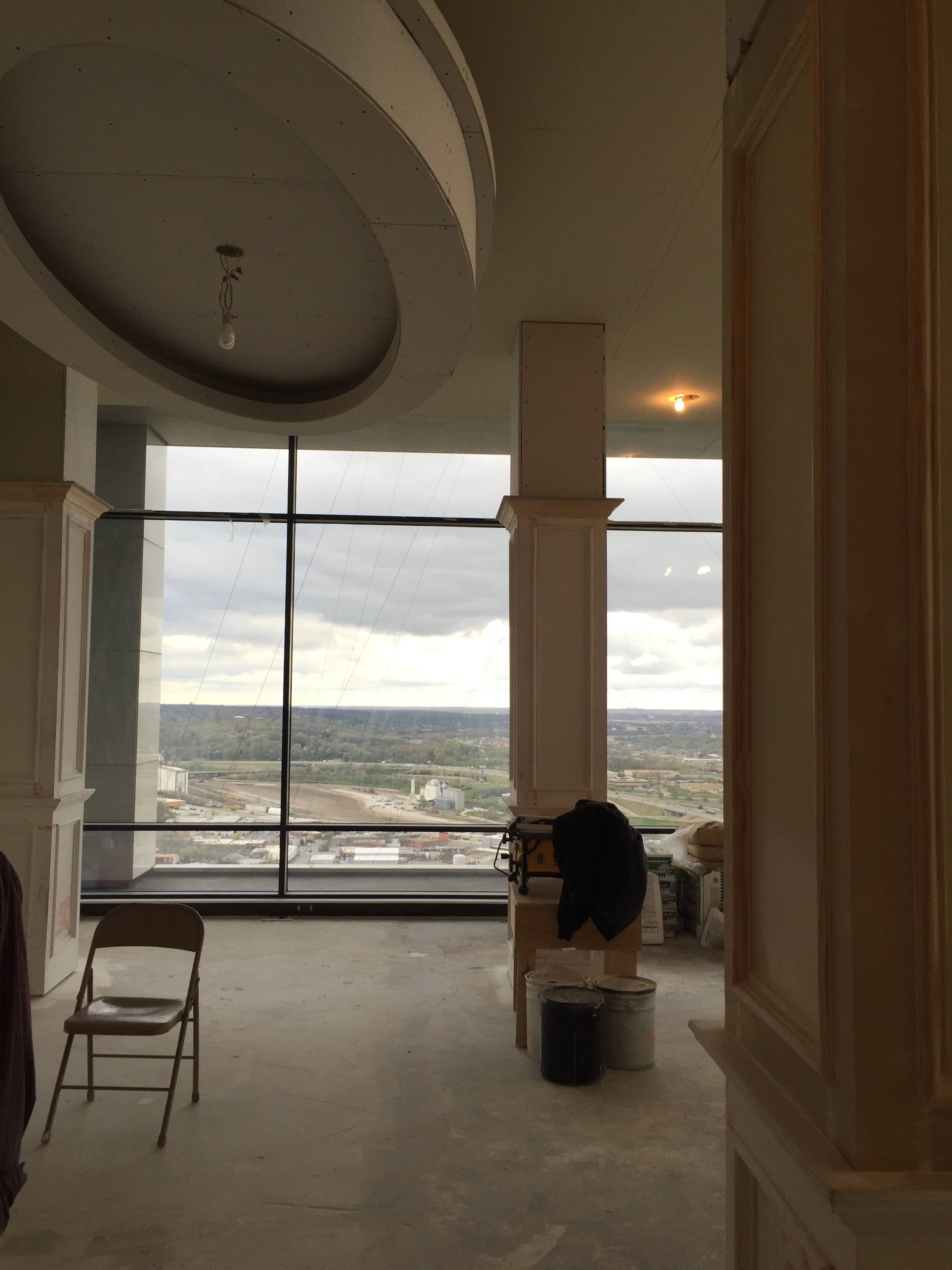 An in-progress interior design project featuring a partially constructed room with a large oval recessed ceiling. The space includes tall columns and large floor-to-ceiling windows offering expansive views of the cityscape below. The room is currently unfinished, with exposed floors and walls, construction materials, and equipment visible. A lone folding chair sits in the center of the space, indicating ongoing work and preparation for further design elements to be added. The overcast sky outside enhances the raw and developing nature of the project.
