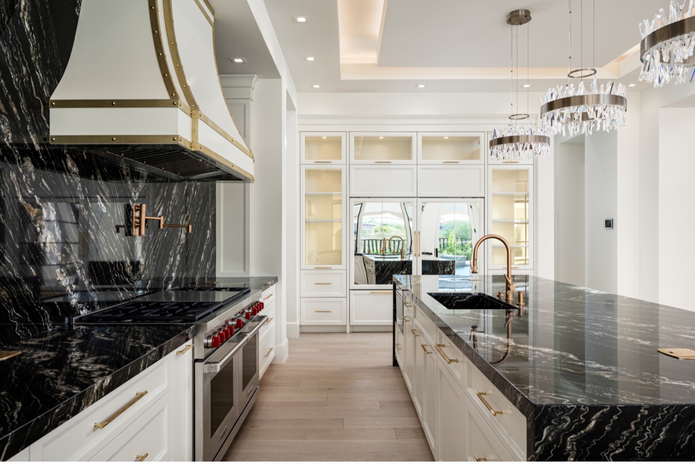 A luxurious, modern kitchen featuring sleek black marble countertops with striking white veining. The kitchen is equipped with high-end stainless steel appliances, including a professional-grade stove with red knobs and a stylish range hood adorned with gold accents. The island includes a deep sink with a gold faucet, complemented by contemporary pendant lights with crystal detailing that hang above, adding elegance and sparkle to the space. The cabinetry is white with gold handles, and a built-in refrigerator blends seamlessly with the cabinets. The room's design is enhanced by a tray ceiling with recessed lighting, creating a bright and inviting atmosphere.